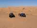 1970 Volkswagen Beetle - Two off-road vehicles, a blue Volkswagen Beetle and a black UTV, are parked in sandy desert dunes under a clear blue sky.