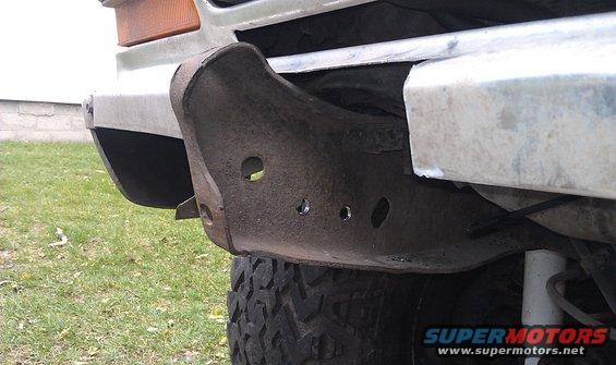 Close-up of a rusty metal front bumper section with holes drilled in it, showing part of a tire and grass