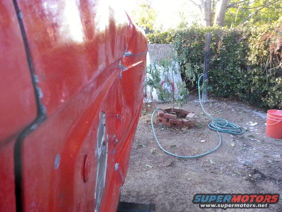 close-up of red vehicle side panel with dents and rust spots, hose and bucket in background
