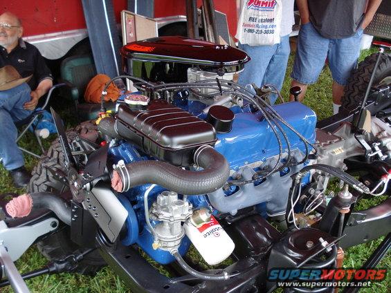 Blue V8 engine with black air cleaner and fan shroud. People and a trailer are in the background