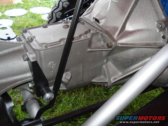 Gray metal transmission housing with black shifter linkage and silver exhaust pipe. Tire tread visible in background