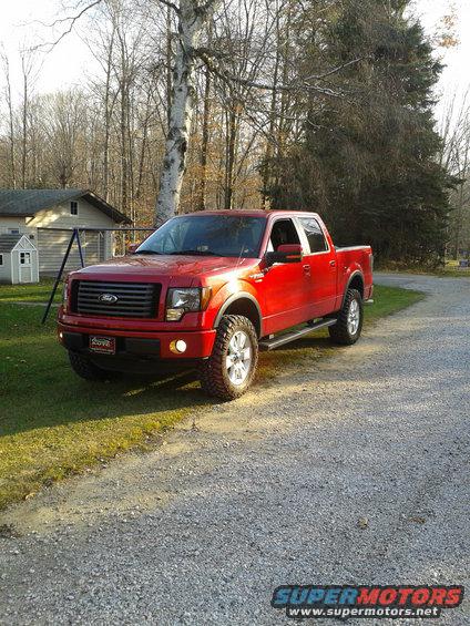 Red pickup truck with aftermarket wheels and tires, parked on a gravel driveway