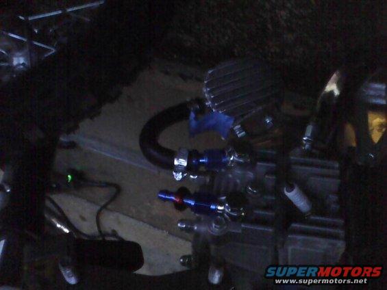Close-up of engine components including hoses, clamps, and what looks like a spark plug. Blue fittings are visible