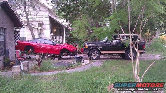 Black and tan Ford Bronco parked on dirt lot next to a red car on blocks