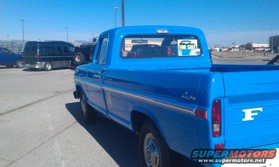 1973 Ford F-100 pickup truck, blue, with a "For Sale" sticker on the rear window. Other vehicles are parked nearby
