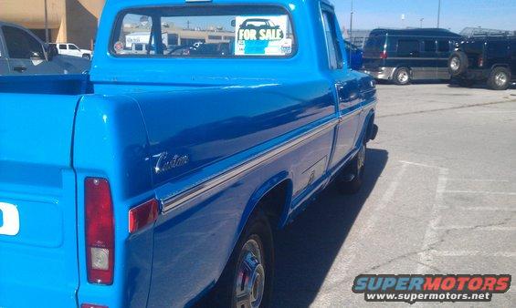 Side view of a blue pickup truck with a chrome trim strip and 'Custom' badging. A 'For Sale' sign is on the rear window