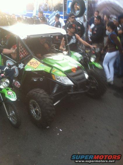 Off-road buggy with dirt bikes next to it, people around