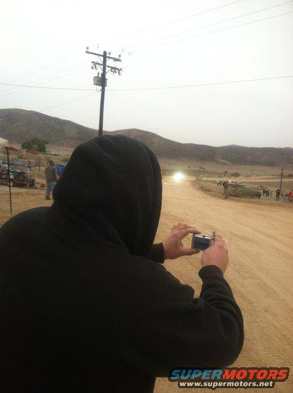 Person in hoodie taking photo of off-road race with hills and power lines in background