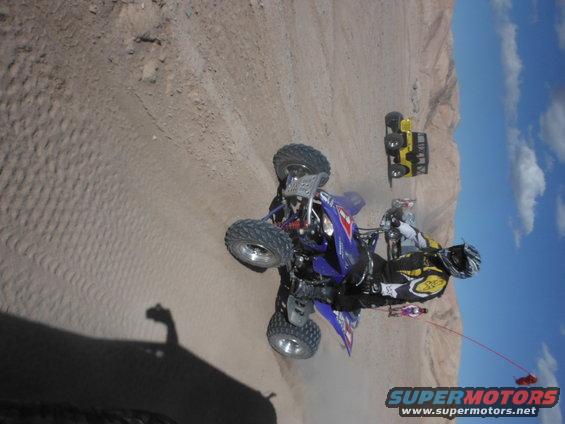 Quad bike with rider in a desert landscape, another yellow vehicle in the background