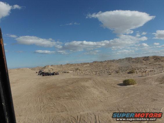 Several off-road vehicles parked in a sandy desert area with hills and a blue sky with clouds