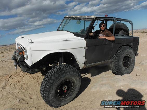 White and black Ford Bronco with large tires and a roll cage, driven by a man in sunglasses