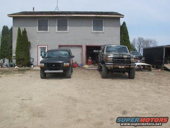 Two SUVs parked in a dirt lot. The one on the right is a dark-colored Ford Bronco with a brush guard