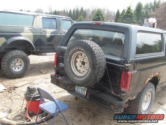 Two Ford Broncos parked on dirt; one is black with a rear spare tire, the other is camouflage. A shop vac and chair are nearby