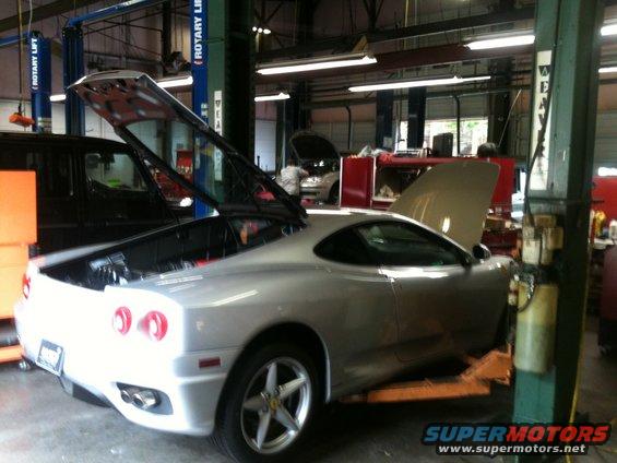 Silver sports car with rear hood open, on a lift in a garage. Another black vehicle is to the left