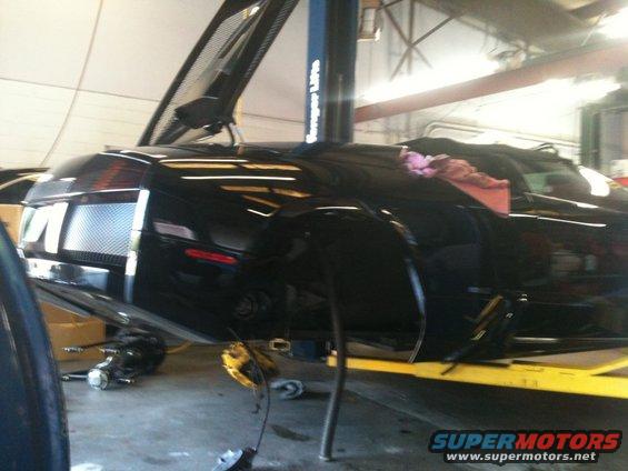Black sports car with hood open on a lift, front wheel removed. Supermotors.net logo visible