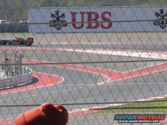 Red and black racing car on a track, seen through a fence