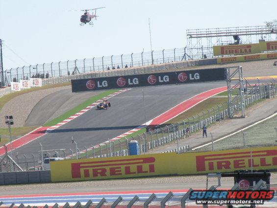 Race track with a single-seater race car on the track and a helicopter in the sky. LG and Pirelli banners are visible