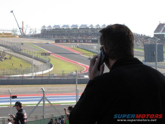 Race track with grandstands full of spectators and a man in the foreground talking on a cell phone
