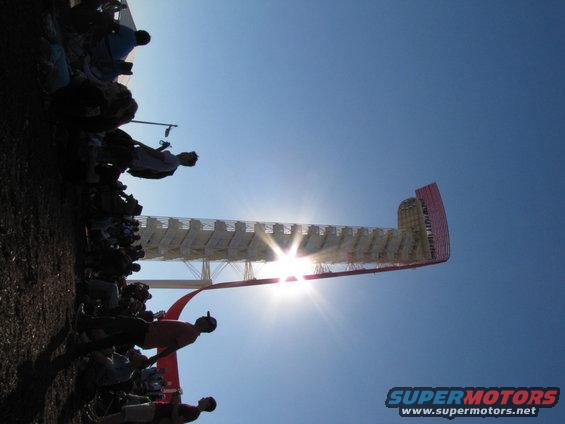 Crowd silhouetted against the sun at a race track with a large structure in the background