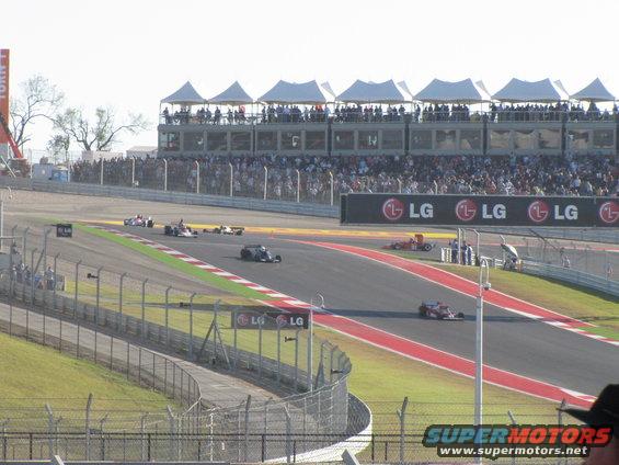 Formula 1 race cars on a track with grandstands and spectators. LG logos visible on a banner