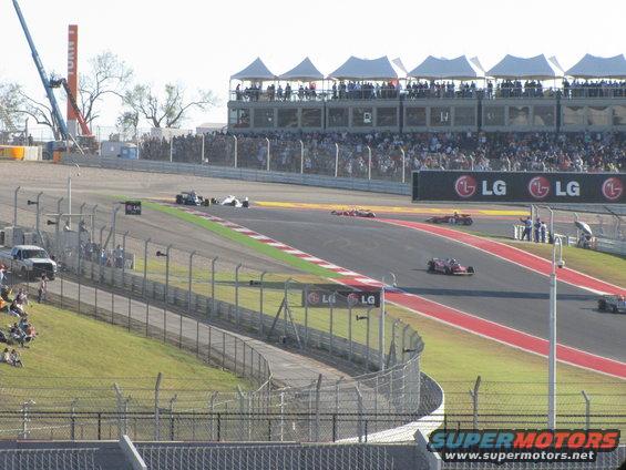 Race cars on a track with spectators in grandstands and a crane in the background