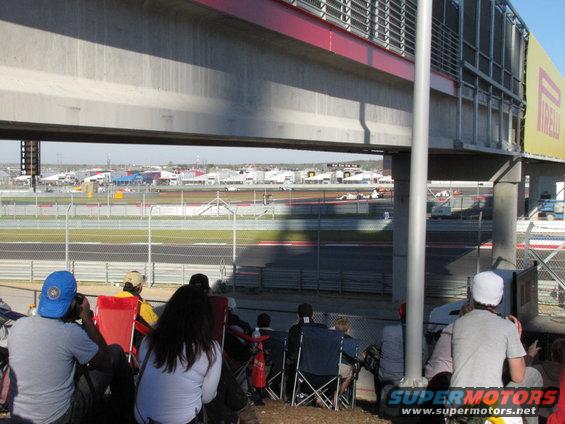 Spectators watch race cars on a track from under a concrete overpass. Buildings and a yellow Pirelli banner are visible in the background
