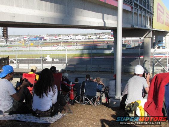 Race cars on a track viewed from under a grandstand with spectators sitting on the ground