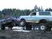1988 Ford Bronco - Two heavily modified trucks tackle a muddy, snowy obstacle course in a wooded area.
