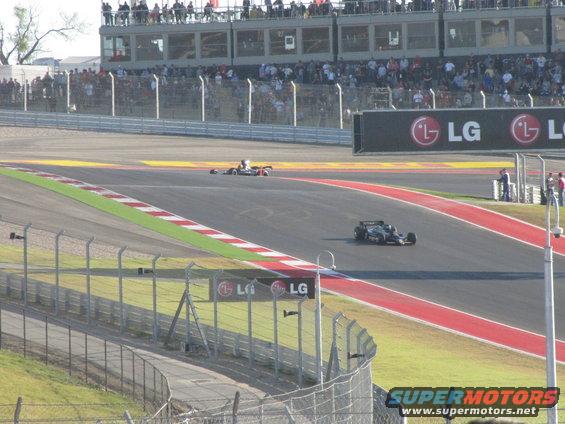 Two formula 1 cars race on a track with spectators in the background and LG billboards