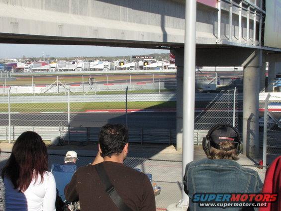 Spectators watch a race on a track, with buildings and trucks in the background