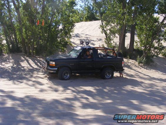 Black 1996 Ford Bronco with roll cage driving on sand dunes near trees