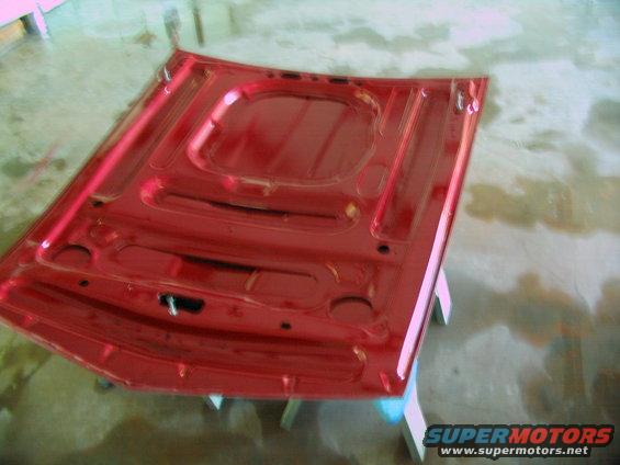 Red car hood underside, viewed from above, showing structural ribs and mounting points. Supported by white stands