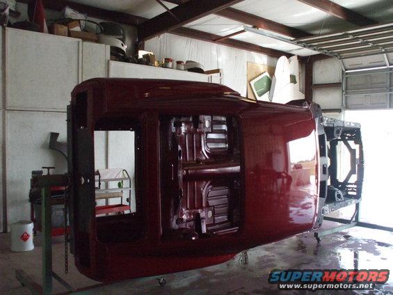 Maroon car body upside down on a metal frame, showing the underside and interior, in a workshop