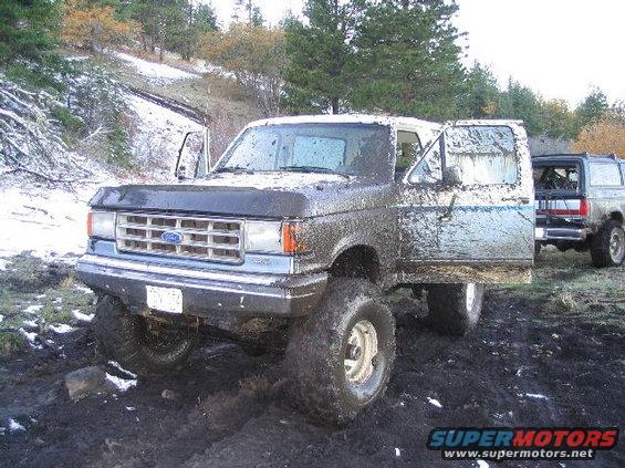 Muddy Ford Bronco with doors open, another Bronco behind it, in a snowy, muddy area