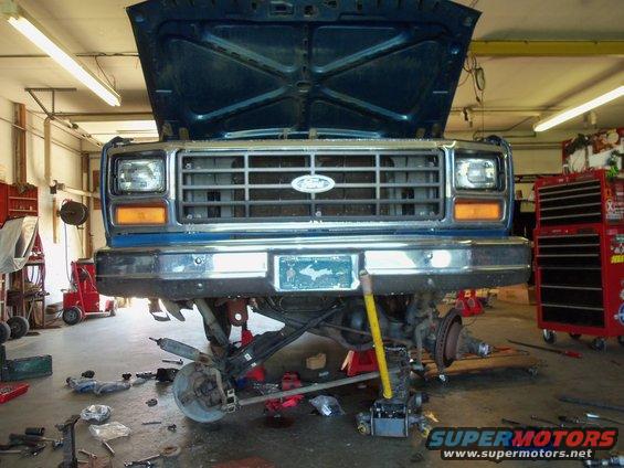 Front of a blue Ford truck with hood up, undergoing front-end work. Tools and parts are scattered on the garage floor