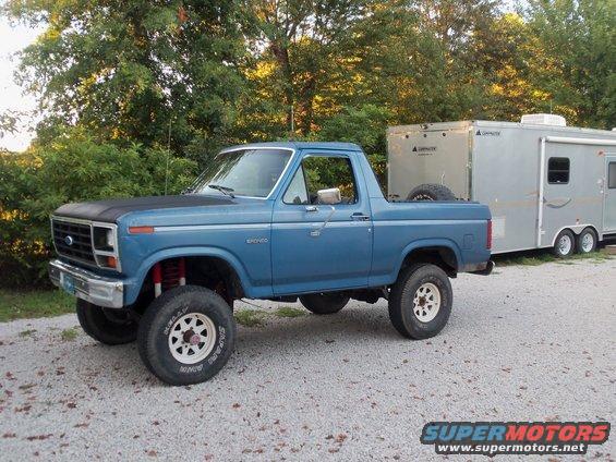 Blue Ford Bronco with lifted suspension, towing a silver trailer. The Bronco has a black hood accent and white wheels