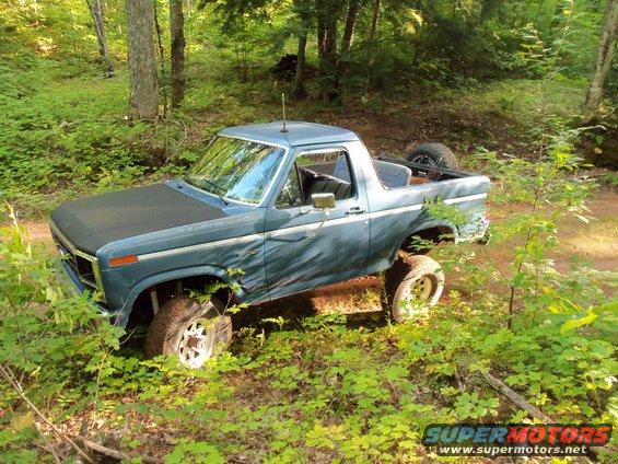Blue Ford Bronco with a black hood, lifted suspension, and spare tire in the bed, parked off-road