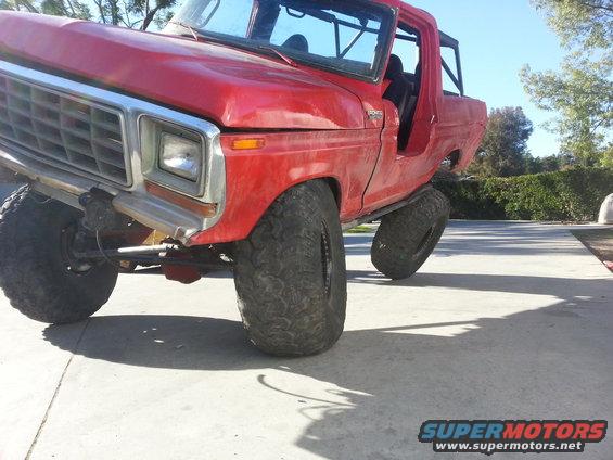 Red 1978 Ford Bronco with large off-road tires, front driver side tire angled up, driver door removed