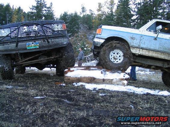Two muddy off-road trucks, one black and one blue and white, are positioned on a log