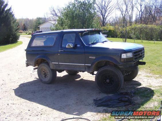 Dark blue Ford Bronco with aftermarket wheels and a sun visor. It's parked on a dirt driveway