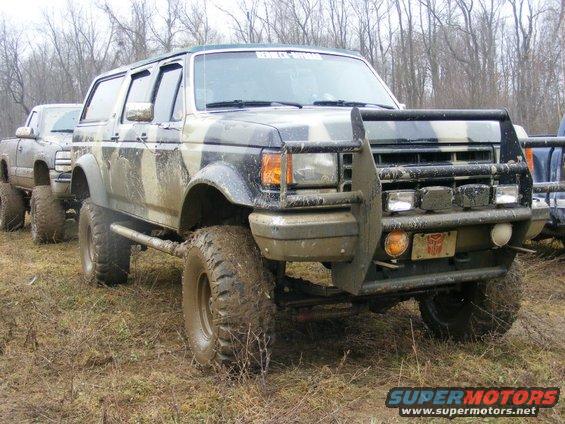 Muddy camouflage 1992 Ford Bronco with aftermarket bull bar and off-road tires, parked next to another lifted truck