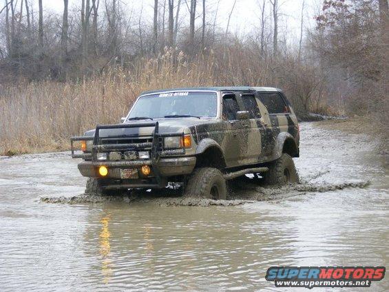 Muddy SUV driving through water, has a brush guard and camouflage paint