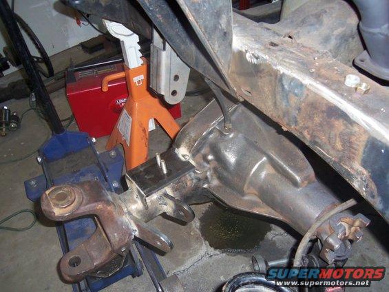 Close-up of a vehicle's rear axle and frame, supported by jacks and a blue hydraulic lift. A red Lincoln welder is in the background