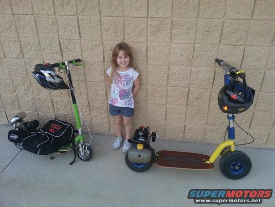 Young girl standing between two motorized scooters, one green and one yellow, against a brick wall