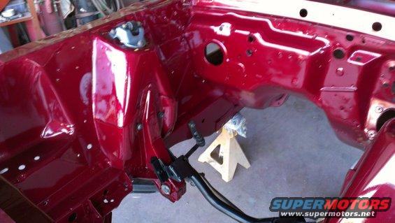 Red painted engine bay frame with visible suspension mounting points and holes, supported by a wooden jack stand