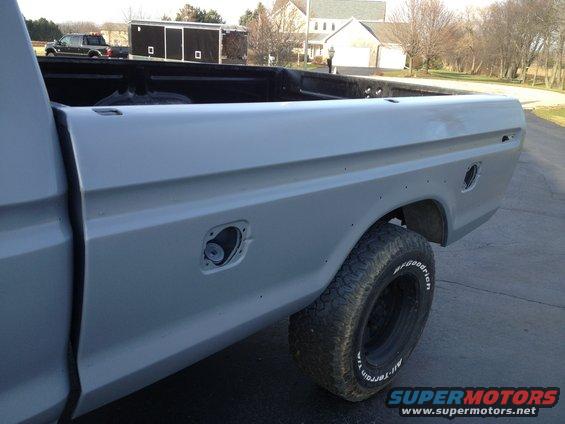 Side view of a gray pickup truck bed with a fuel door and a tire. Another truck and trailer are in the background
