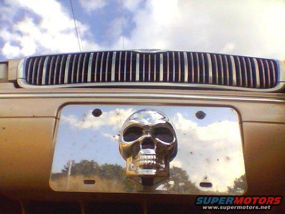 Chrome skull on license plate frame in front of vertical bar grille. The car is gold