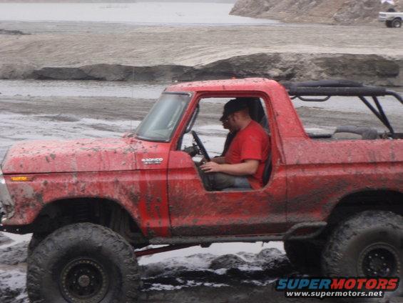 Red 1978 Ford Bronco with a roll cage, covered in mud, driving off-road
