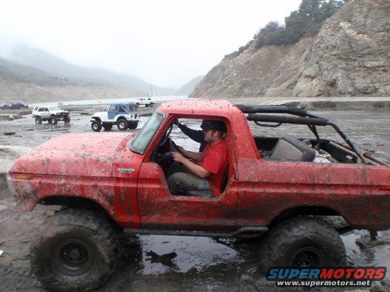 Red 1978 Ford Bronco driving through mud with other off-road vehicles in background
