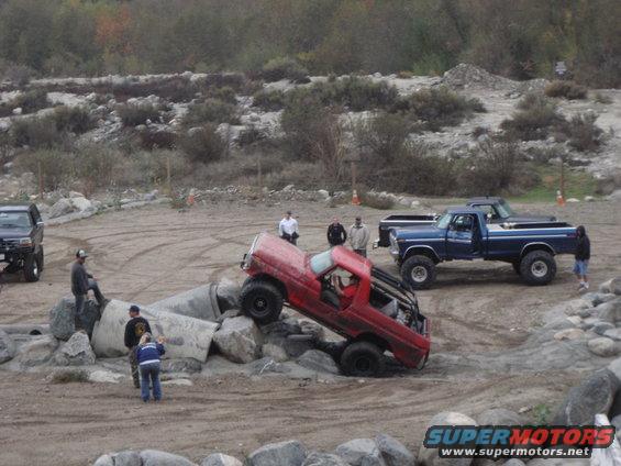 Red pickup truck with roll cage attempting an obstacle course, blue pickup truck parked nearby